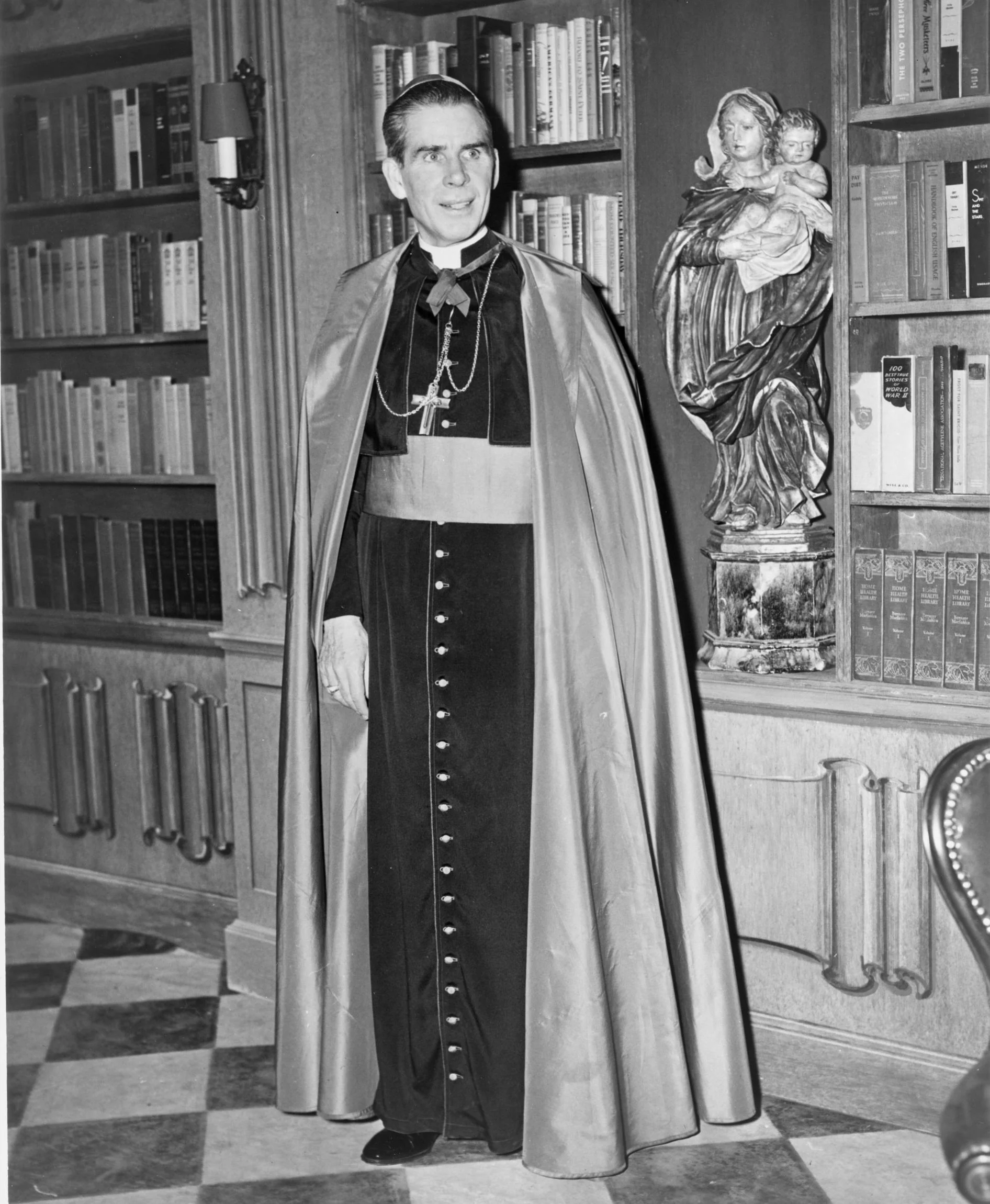 Fulton J. Sheen im Jahr 1952, stehend neben einer Madonna-mit-Kind-Statue und Bücherregalen
